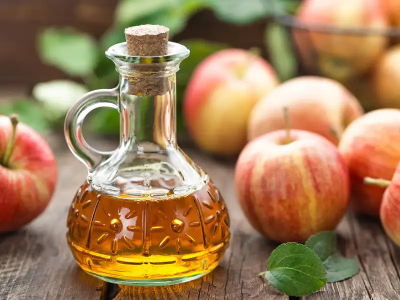 Vinagre de manzana en un dispensador y manzanas al fondo sobre una mesa.